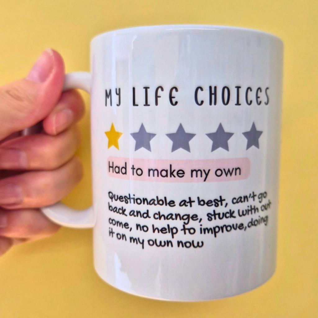 A hand holding a white ceramic mug with a humorous quote that reads 'MY LIFE CHOICES' above a one-star rating, with additional text below, set against a solid yellow background.