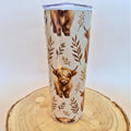 A tall insulated tumbler with a clear lid, decorated with a pattern of Highland cows and brown leaves on a cream background, displayed on a round wood slice against a light gray background.