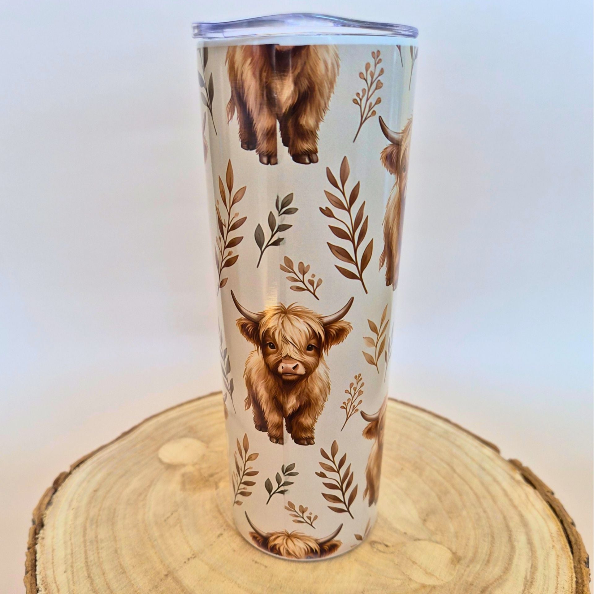 A tall insulated tumbler with a clear lid, decorated with a pattern of Highland cows and brown leaves on a cream background, displayed on a round wood slice against a light gray background.