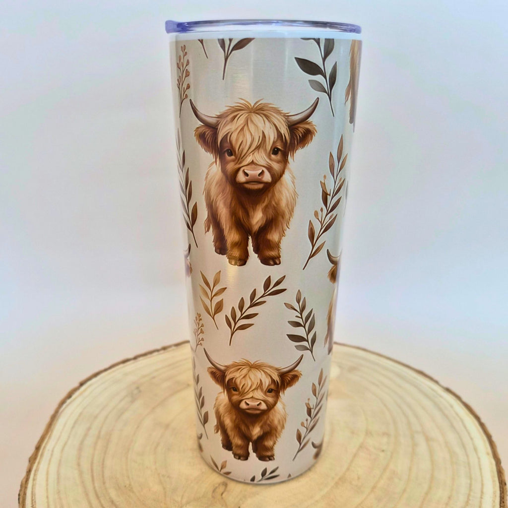 A tall insulated tumbler with a clear lid, decorated with two brown Highland cow illustrations and leafy botanical motifs on a light background, displayed on a round wood slice against a plain light backdrop.