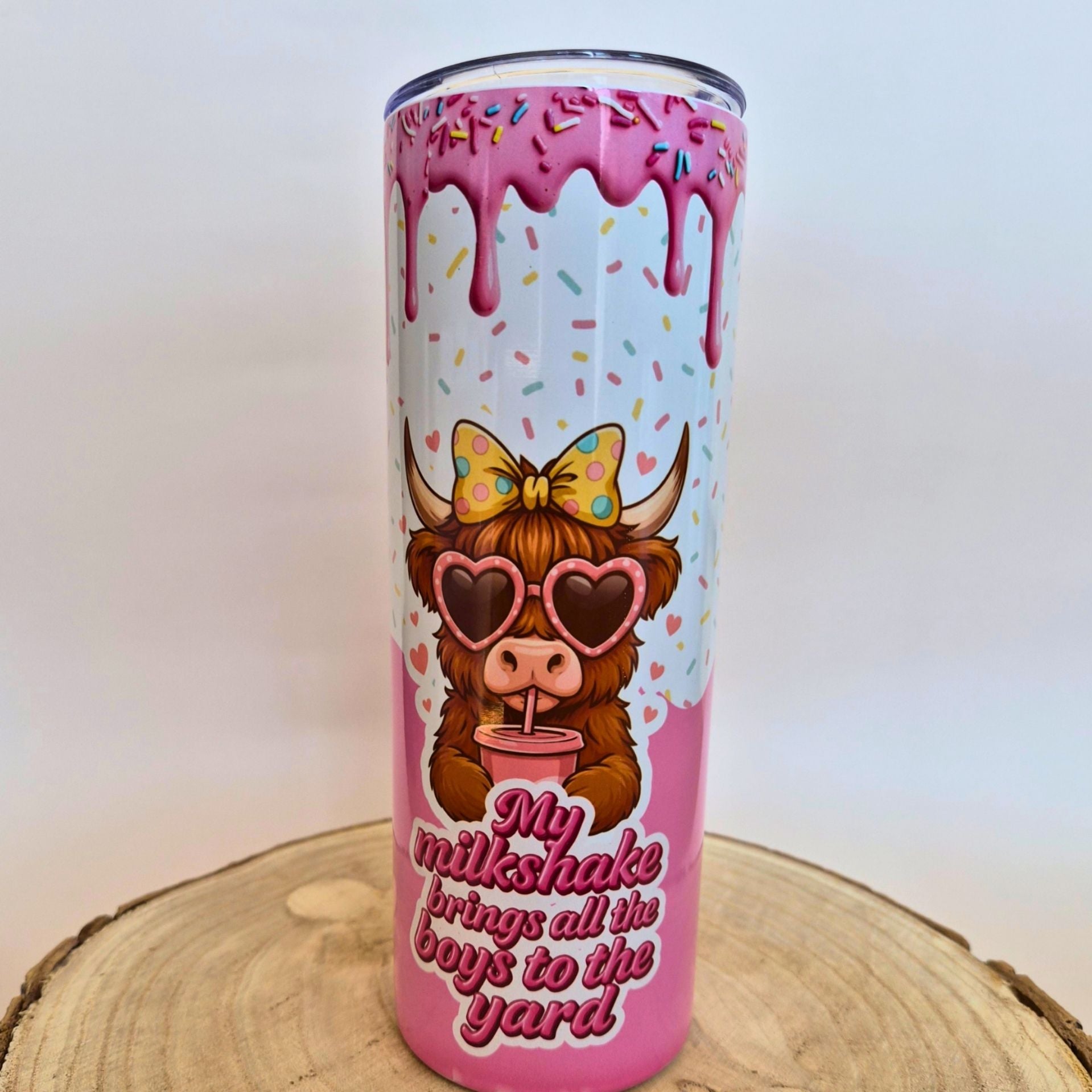 A tall insulated tumbler with a pink and white drip-frosted design, a cartoon cow wearing heart-shaped sunglasses and a bow, and the phrase 'My milkshake brings all the boys to the yard' on a wooden slice background.