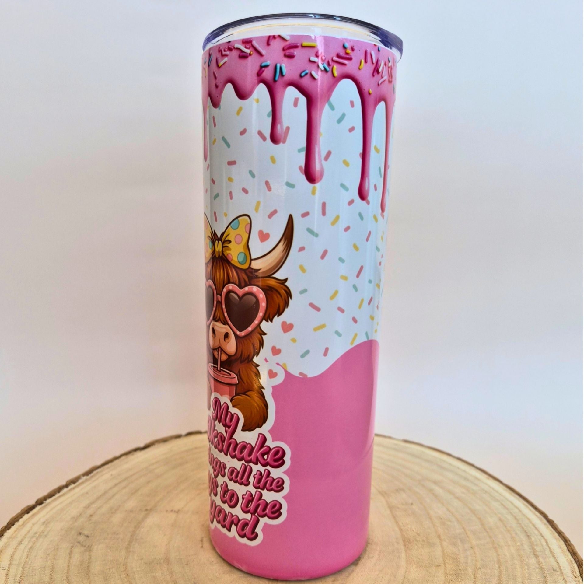 A tall insulated tumbler with a donut-inspired design, featuring pink dripping frosting, colorful sprinkles, a cartoon cow wearing heart-shaped sunglasses, and the phrase 'My shake is all the joy to the yard' on a light wood slice background.