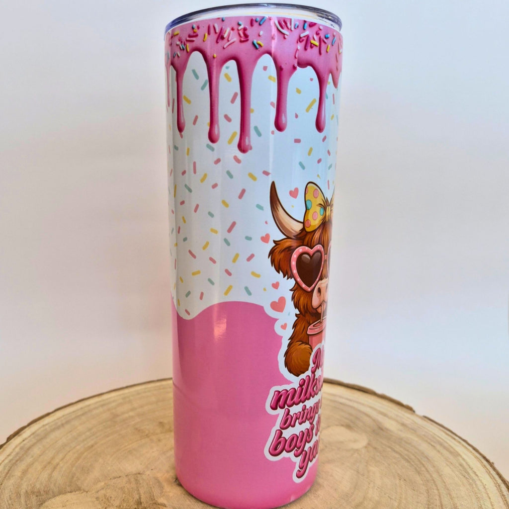 A tall insulated tumbler with a pink and white donut-themed design, featuring dripping pink glaze, colorful sprinkles, a cartoon cow wearing heart-shaped sunglasses, and partial text on a light wood slice background.