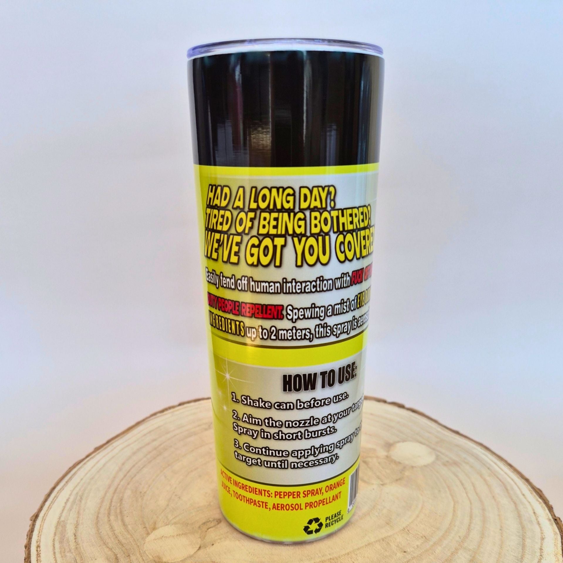 A novelty pepper spray can with a black top and a bright yellow label featuring humorous text and usage instructions, displayed on a circular wood slice against a light gray background.