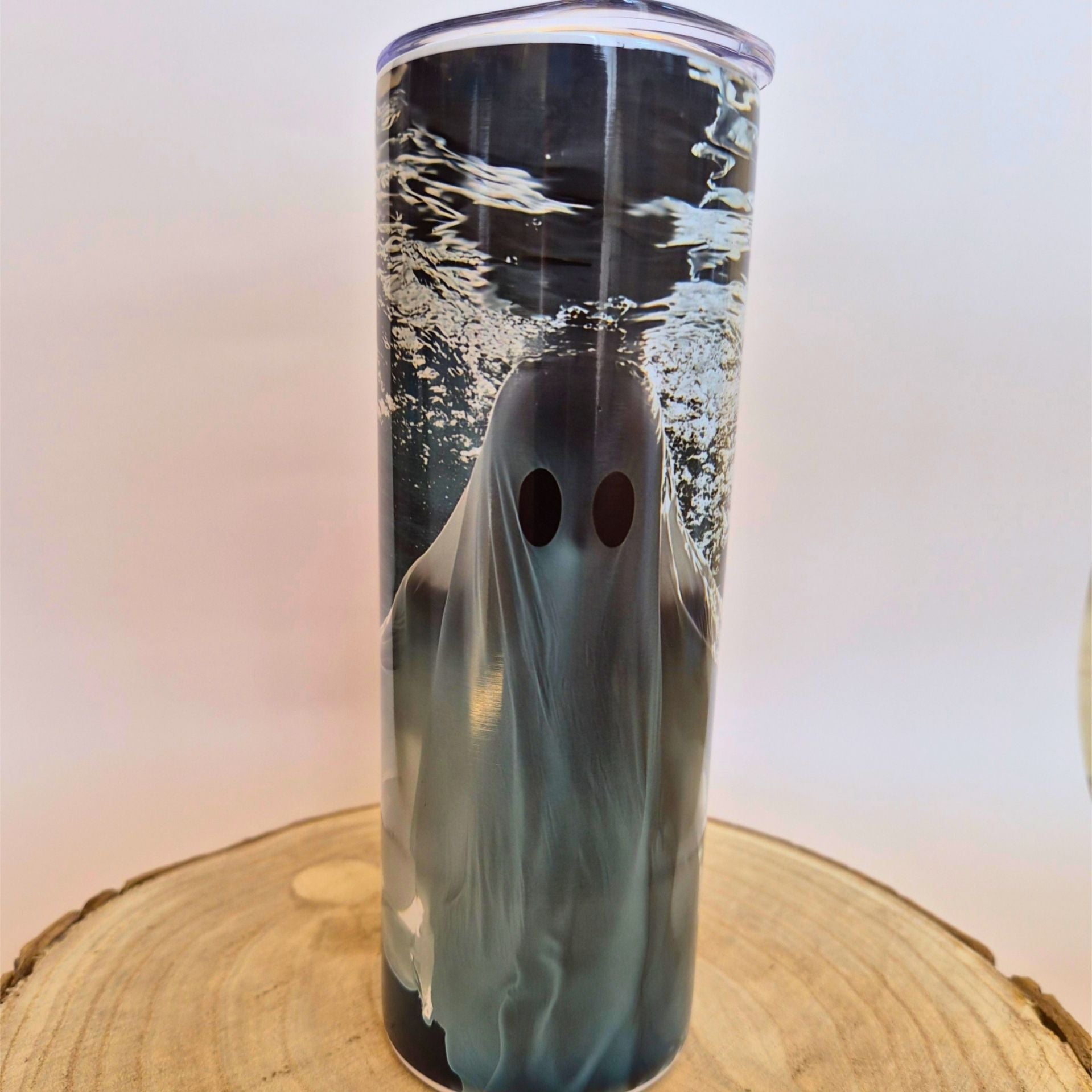 A clear cylindrical tumbler with a printed ghost design under rippling water, shown standing on a round wood slice against a plain light background.