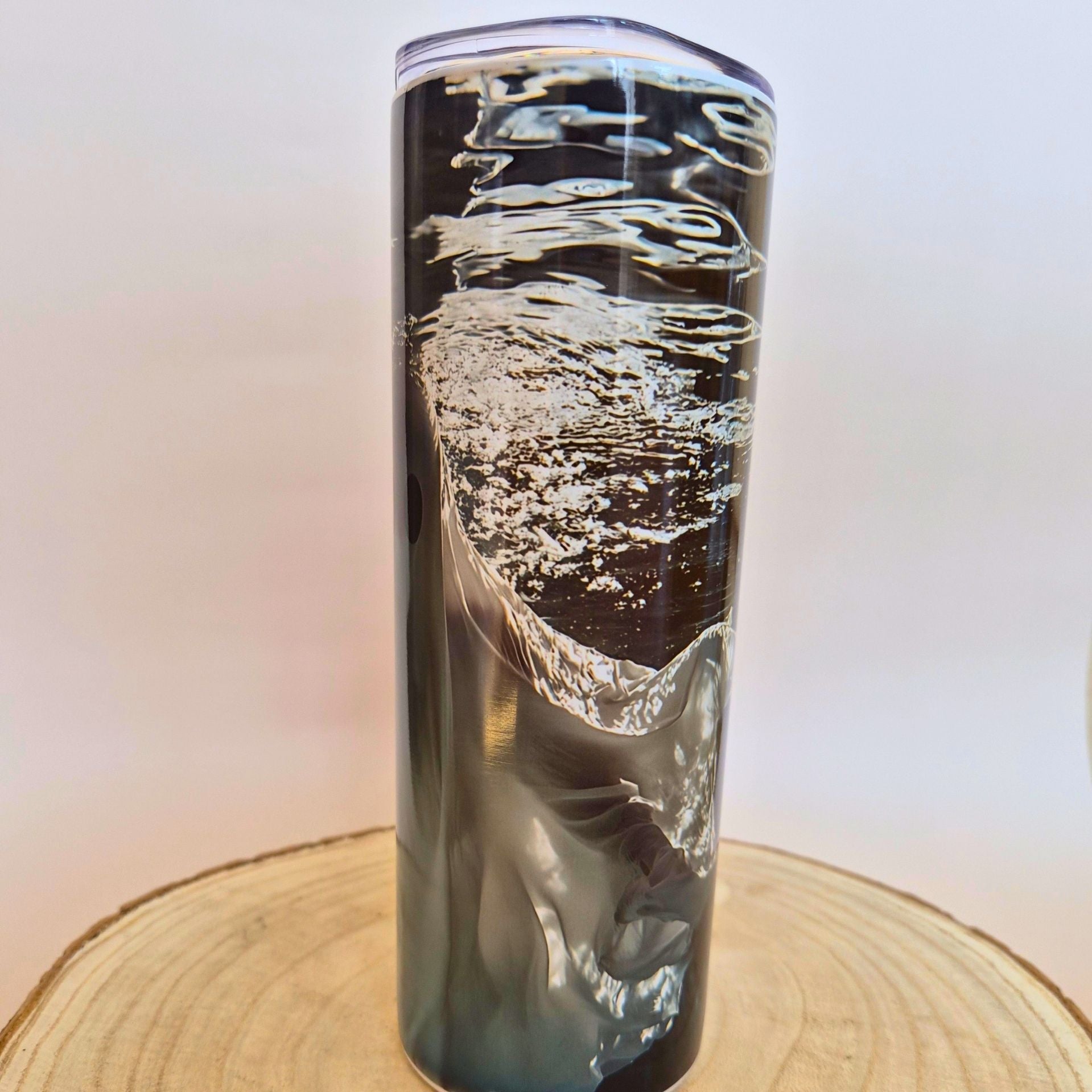 A tall, slim clear tumbler with a printed or embedded wave-like pattern in black, gray, and white, shown standing on a round wood slice against a white backdrop with repeated Memory Metal watermarks.