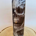 A tall cylindrical tumbler with a detailed black, white, and gray skull design integrated with swirling smoke or flame patterns, displayed on a wooden slice coaster against a neutral light background.