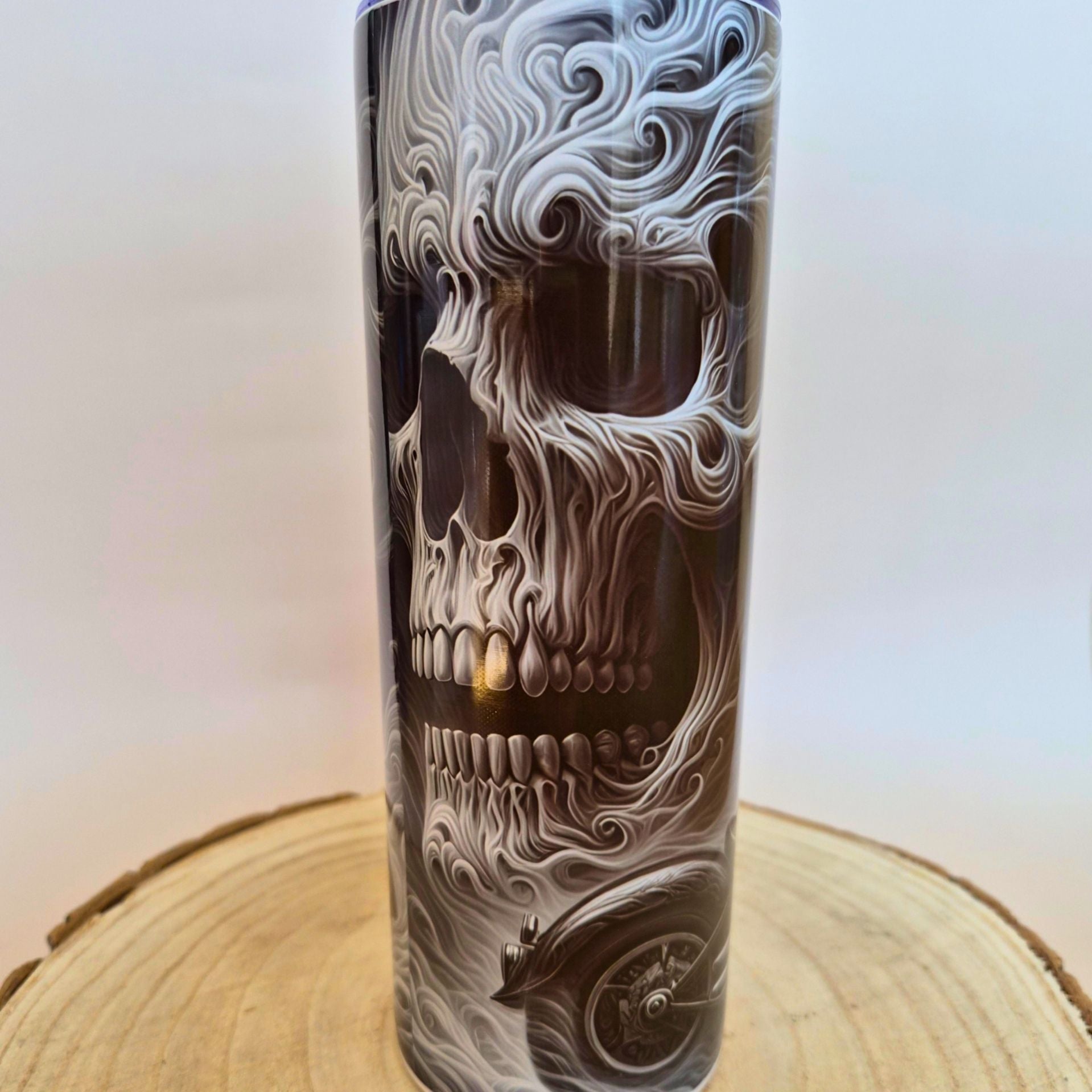 A tall cylindrical tumbler with a detailed black, white, and gray skull design integrated with swirling smoke or flame patterns, displayed on a wooden slice coaster against a neutral light background.