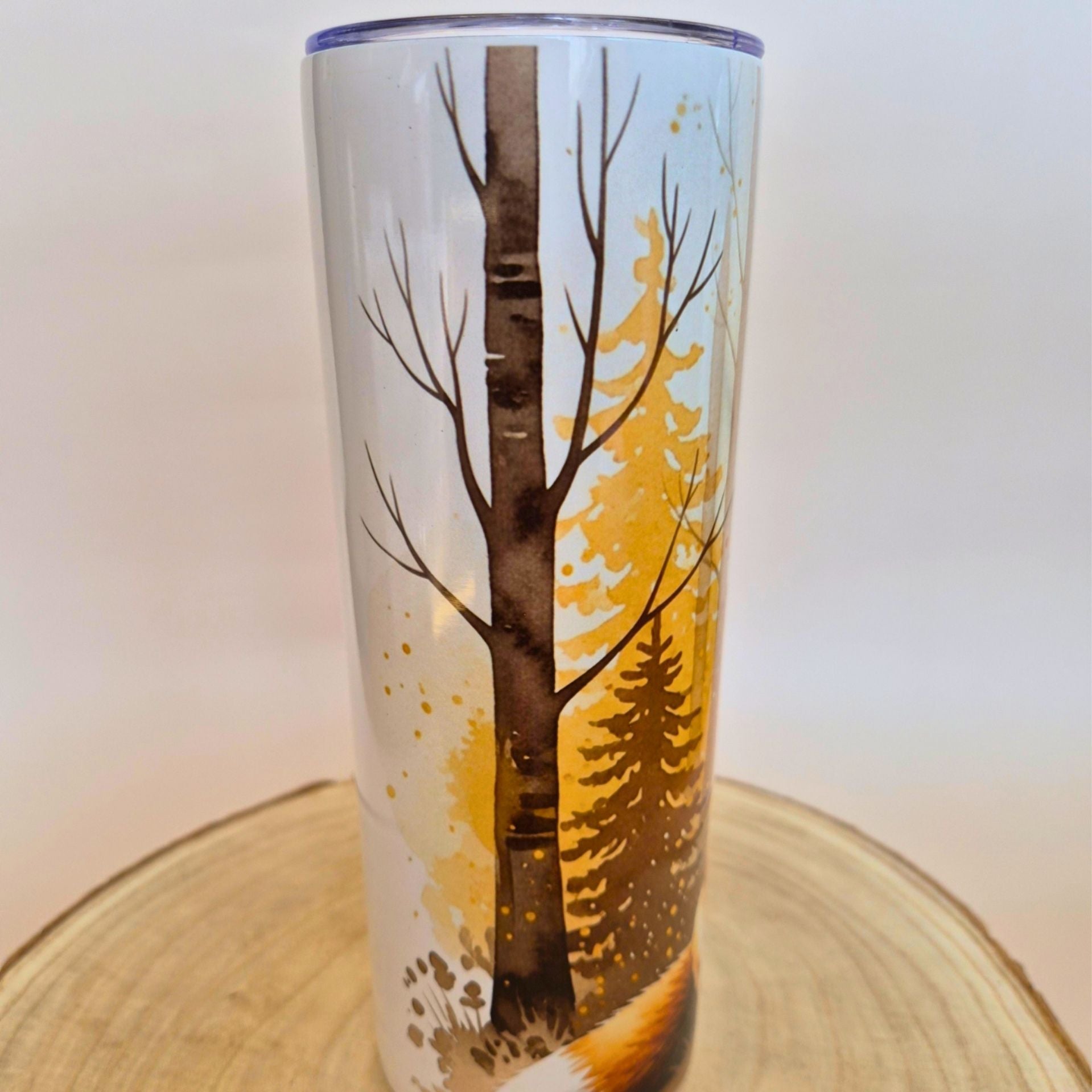 A tall insulated tumbler with a clear winter forest design, featuring a birch tree, pine trees, and a fox, displayed on a round wooden slice against a neutral background.