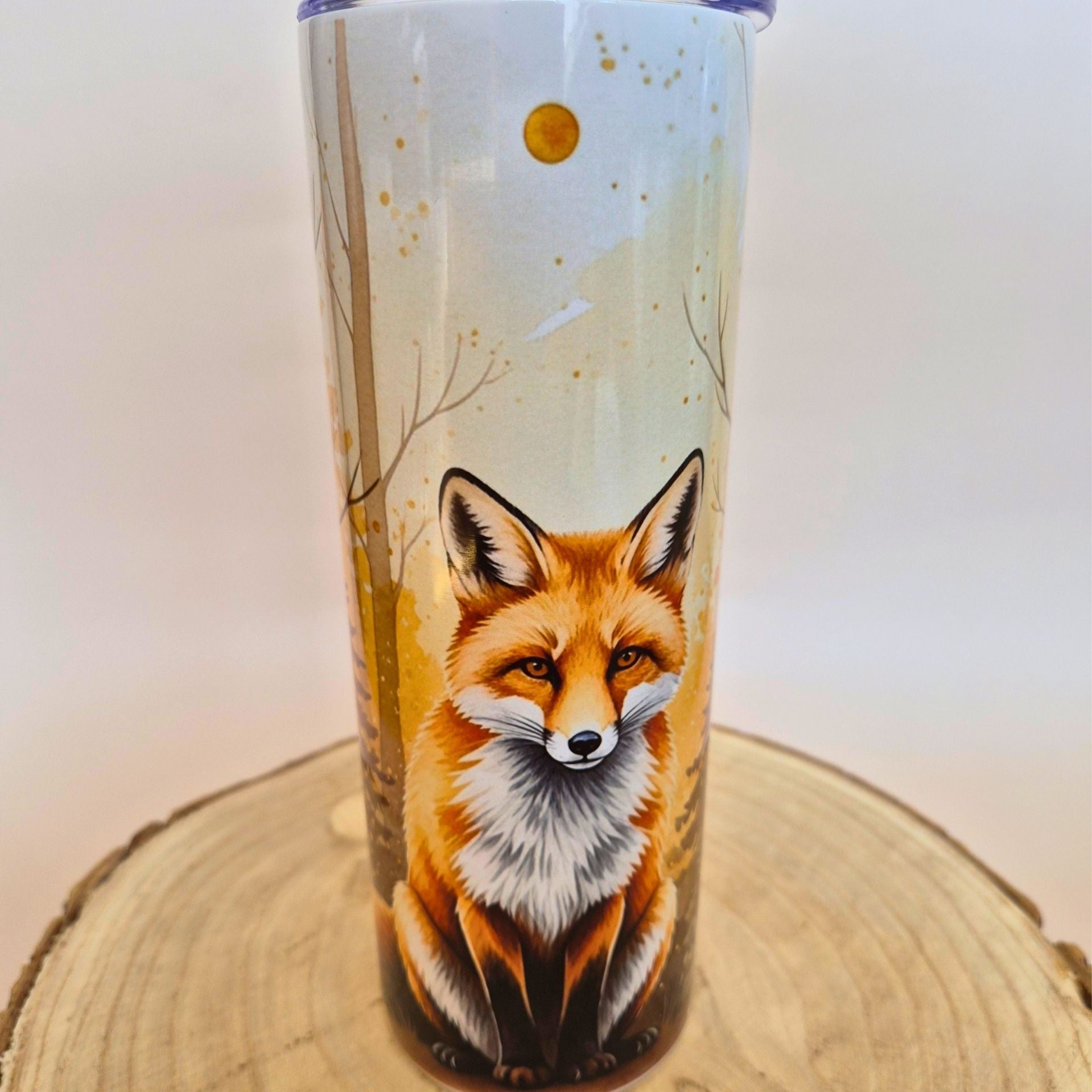 A tall insulated tumbler with a lid, featuring a detailed illustration of a red fox in a light-colored forest scene, displayed on a round wooden slice against a plain light background.