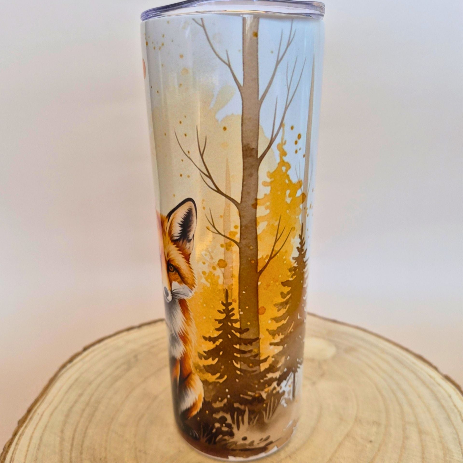 A tall clear tumbler with a printed forest and fox design, shown standing on a round wood slice against a plain light background.
