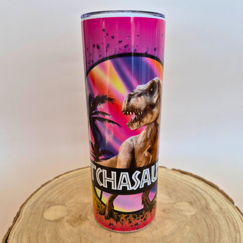 A tall insulated tumbler with a clear lid, featuring a colorful dinosaur graphic and the text 'TCHASAU' on the front, displayed on a round wood slice against a plain light background.