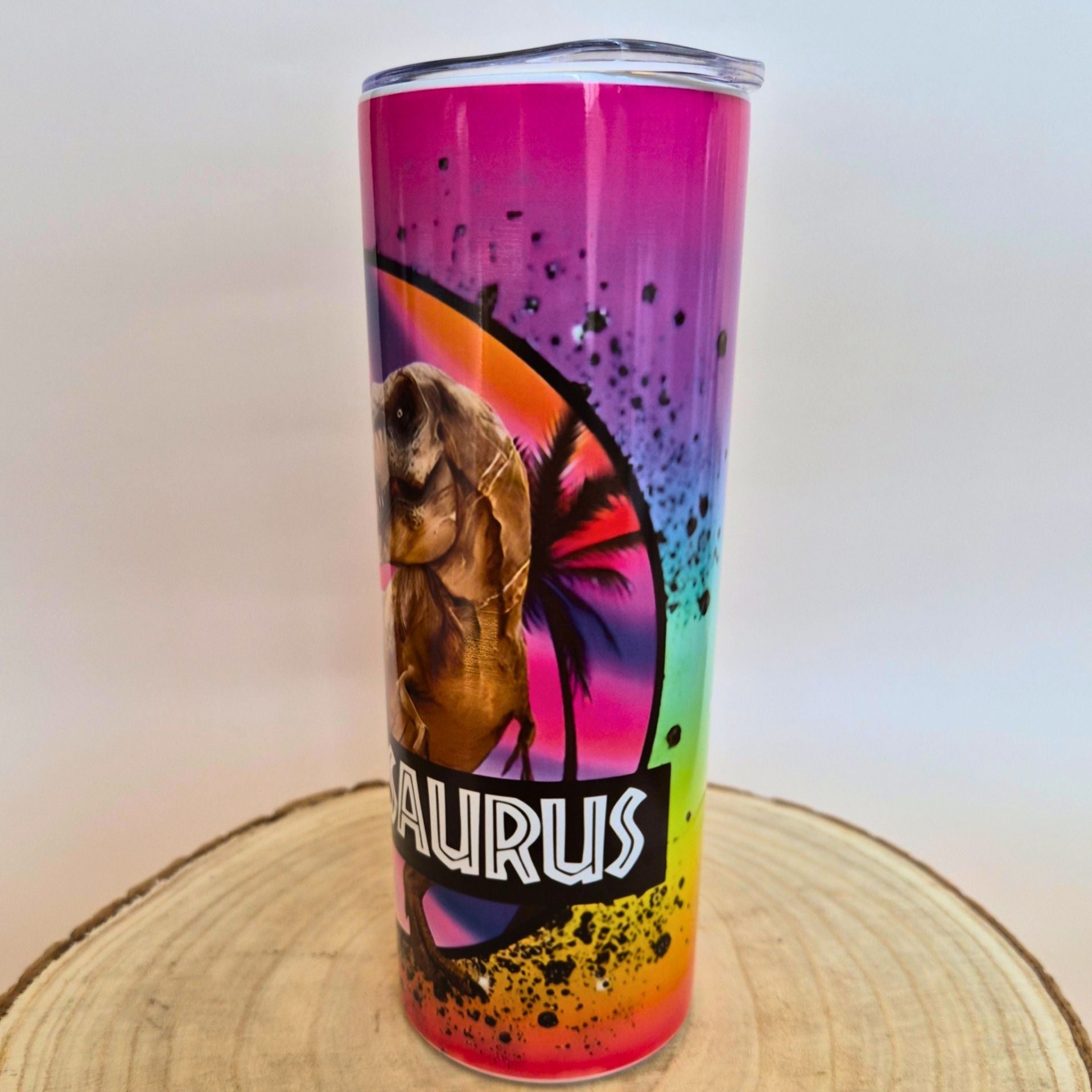 A tall insulated tumbler with a clear lid, featuring a colorful rainbow gradient and a dinosaur illustration with the word 'Taurus' on the front, standing on a round wood slice against a plain light background.