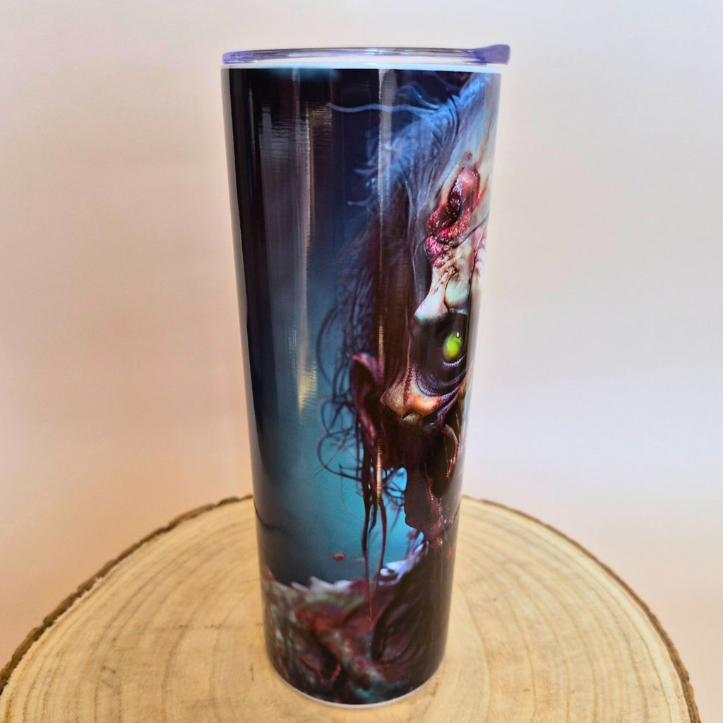 A tall insulated tumbler with a clear lid, featuring a dark fantasy creature illustration in blue, black, purple, and red tones, displayed on a round wood slice against a plain light background.
