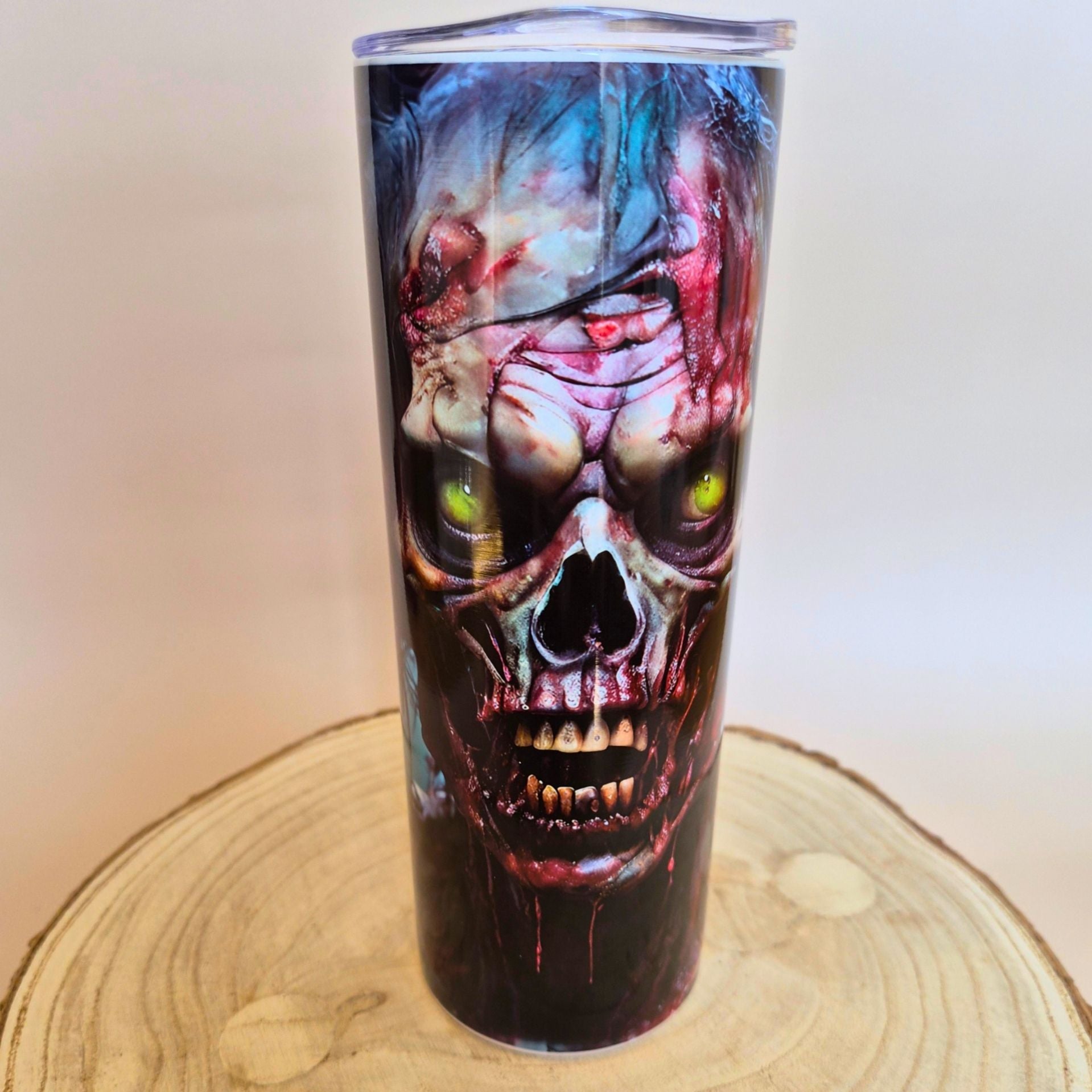 A tall insulated tumbler with a clear lid, featuring a detailed graphic print of a zombie skull with glowing green eyes, displayed on a round wooden slice against a plain beige background.