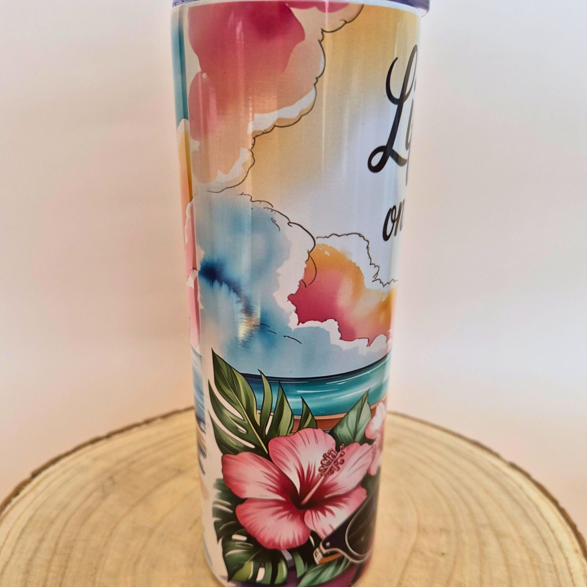 A tall insulated tumbler with a tropical beach and floral design, featuring pink hibiscus flowers, palm leaves, a blue ocean, and a sunset sky. The tumbler is shown standing on a circular wooden slice against a plain light background.