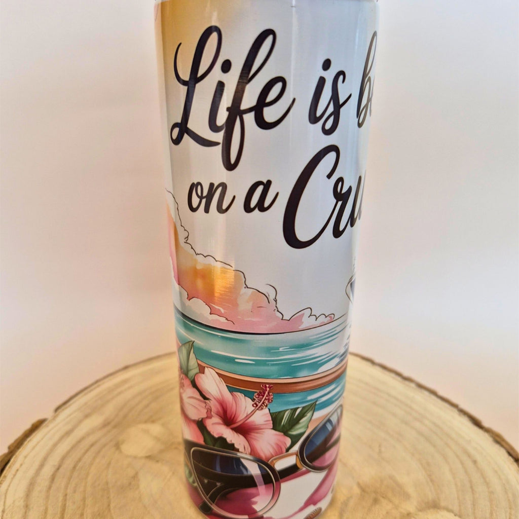 A white insulated tumbler with a tropical beach illustration and the text 'Life is best on a Cruise' in black script, displayed on a round wooden slice against a plain light background.