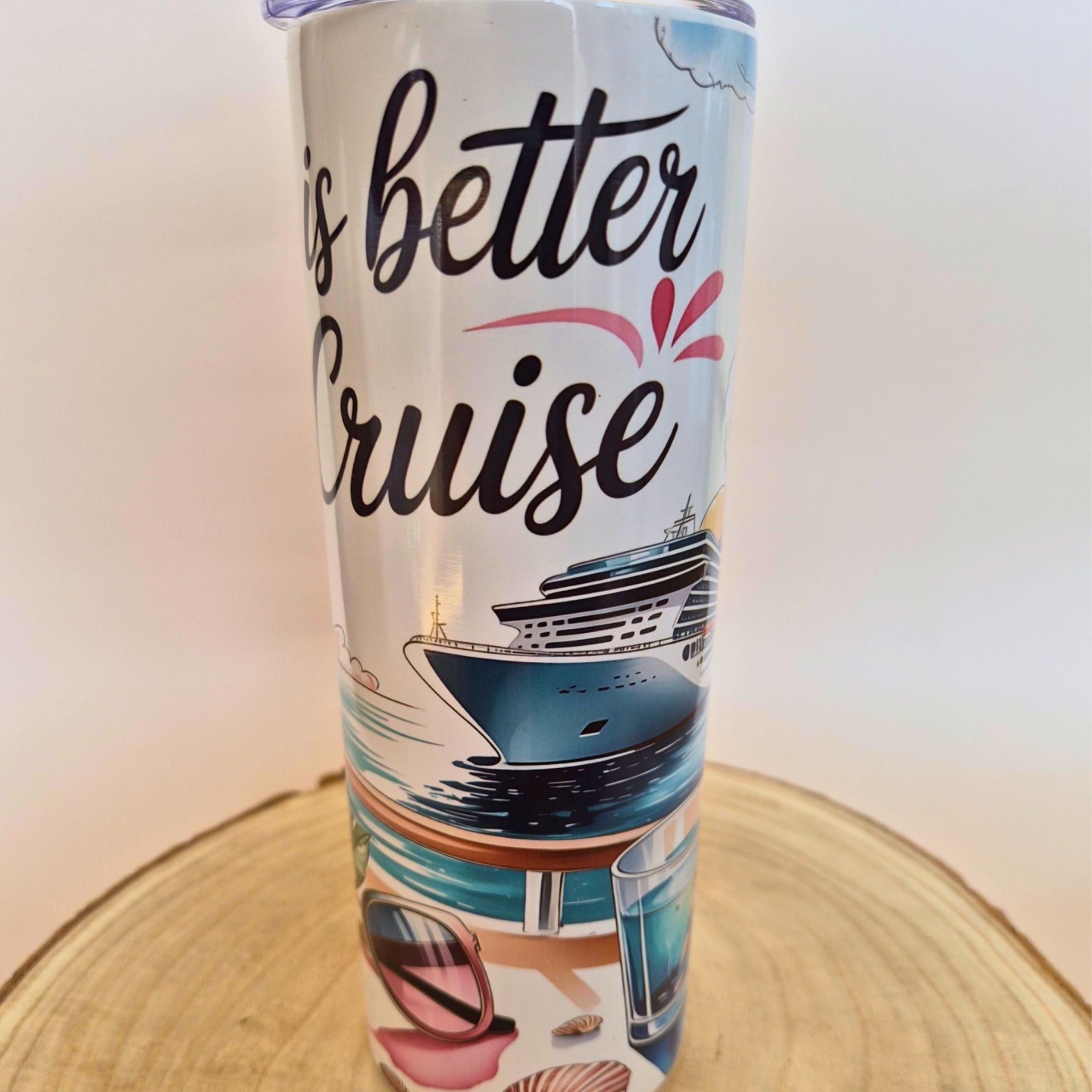 A white insulated tumbler with a travel-themed print that reads 'is better Cruise' in black script, featuring an illustration of a cruise ship, ocean waves, and beach accessories, standing on a round wooden slice against a plain light background.