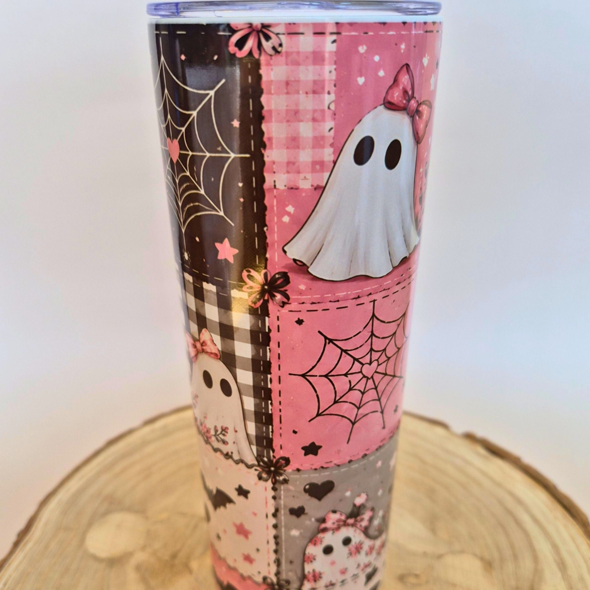A tall insulated tumbler with a Halloween-themed patchwork design, featuring pink gingham, black spiderwebs, white ghosts, and stars, shown on a wooden slice coaster against a white background.