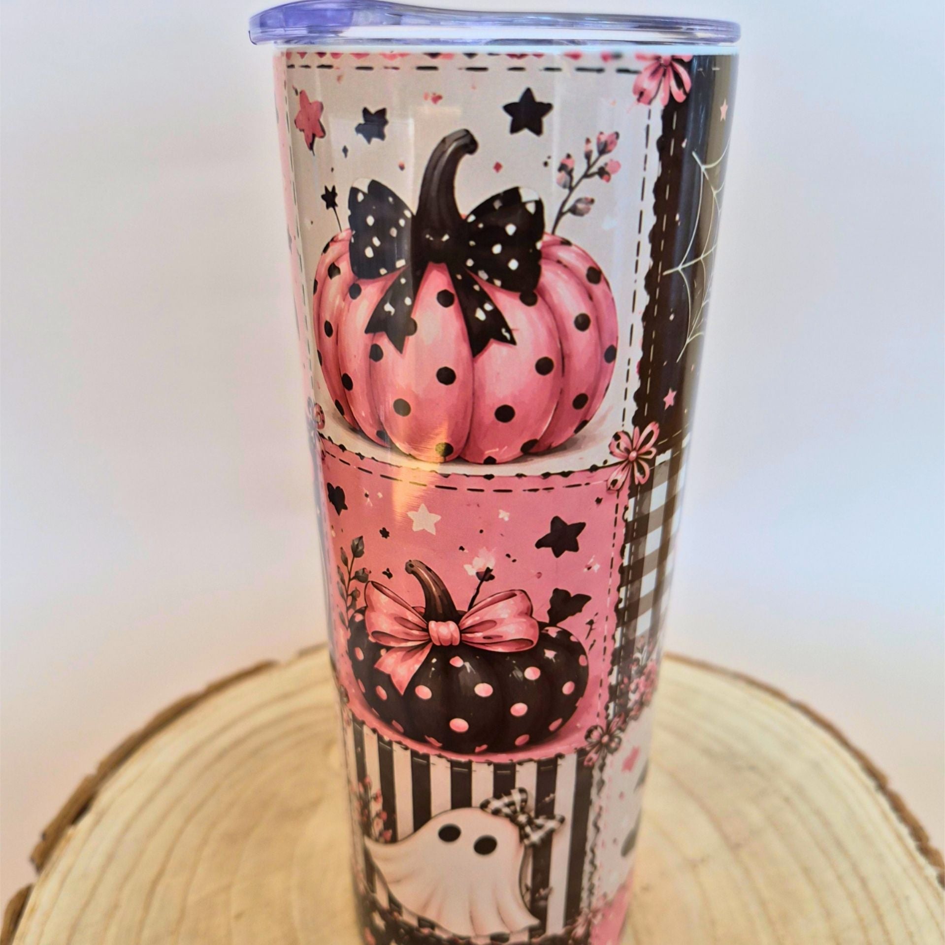 A tall insulated tumbler with a Halloween-themed print featuring pink and black polka-dot pumpkins, black bows, stars, stripes, and a ghost, shown on a wooden slice coaster against a plain light background.