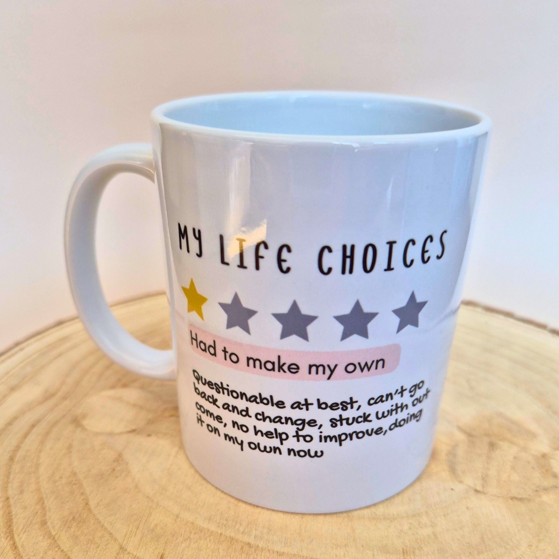 A white ceramic mug with printed text reading 'MY LIFE CHOICES' and a five-star rating graphic, placed on a round wooden surface against a plain light background.