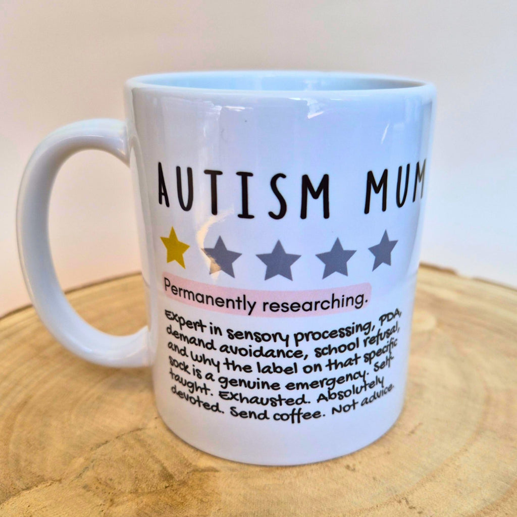 A white ceramic mug with a handle, printed with the text 'AUTISM MUM' and a humorous definition, displayed on a wooden surface against a plain light background.