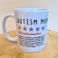 A white ceramic mug with a handle, printed with the text 'AUTISM MUM' and a humorous definition, displayed on a wooden surface against a plain light background.