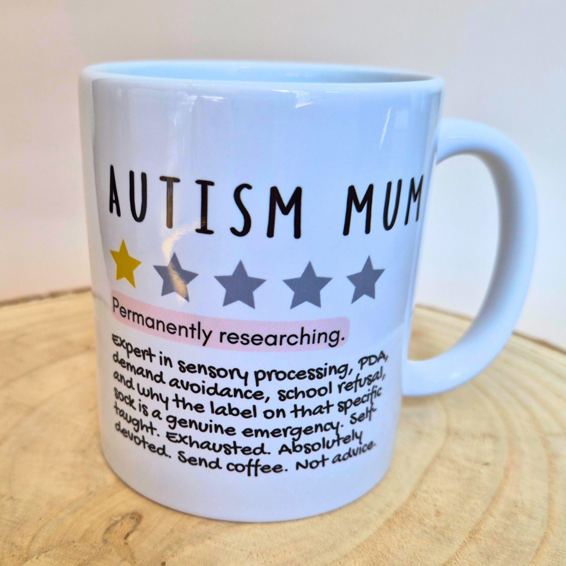 A white ceramic coffee mug with 'AUTISM MUM' printed in black text, accompanied by a row of stars and a humorous definition of autism. The mug is shown on a light wooden surface against a neutral background.